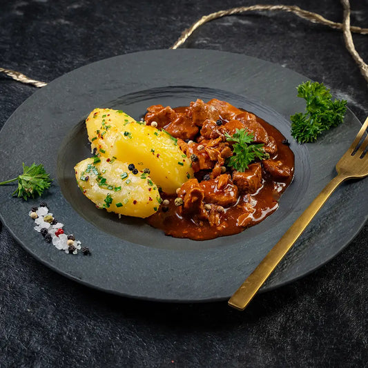 Herzhaftes Gulasch mit würziger roter Sauce, serviert mit Petersilienkartoffeln und garniert mit frischen Kräutern und grobem Pfeffer auf einem dunklen Teller und goldener Gabel auf dunklem Untergrund