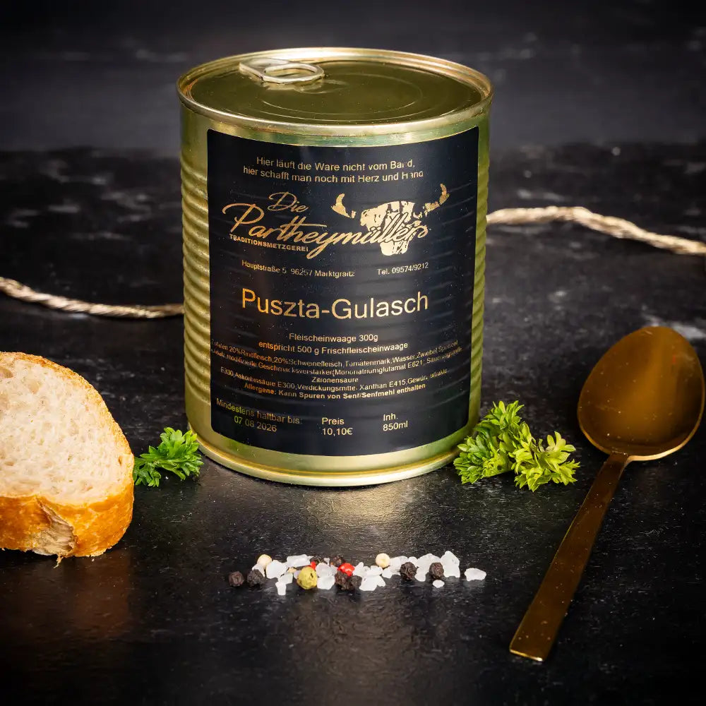 Goldene Konservendose der Metzgerei Partheymüller mit Puszta-Gulasch edel präsentiert auf dunklem Untergrund mit goldenem Löffel, frischen Kräutern, Pfefferkörnern, Meersalz und einer Baguettescheibe.