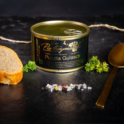 Kleine goldene Konservendose der Metzgerei Partheymüller mit Pusta-Gulasch edel präsentiert auf dunklem Untergrund mit goldenem Löffel, frischen Kräutern, Pfefferkörnern, Meersalz und einer Baguettescheibe.