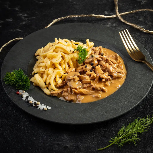 Rahmgeschnetzeltes mit Pilzrahmsauce und Spätzle auf einem schwarzen Teller, garniert mit Petersilie, Pfefferkörnern und Meersalz, daneben eine goldene Gabel auf dunklem Untergrund