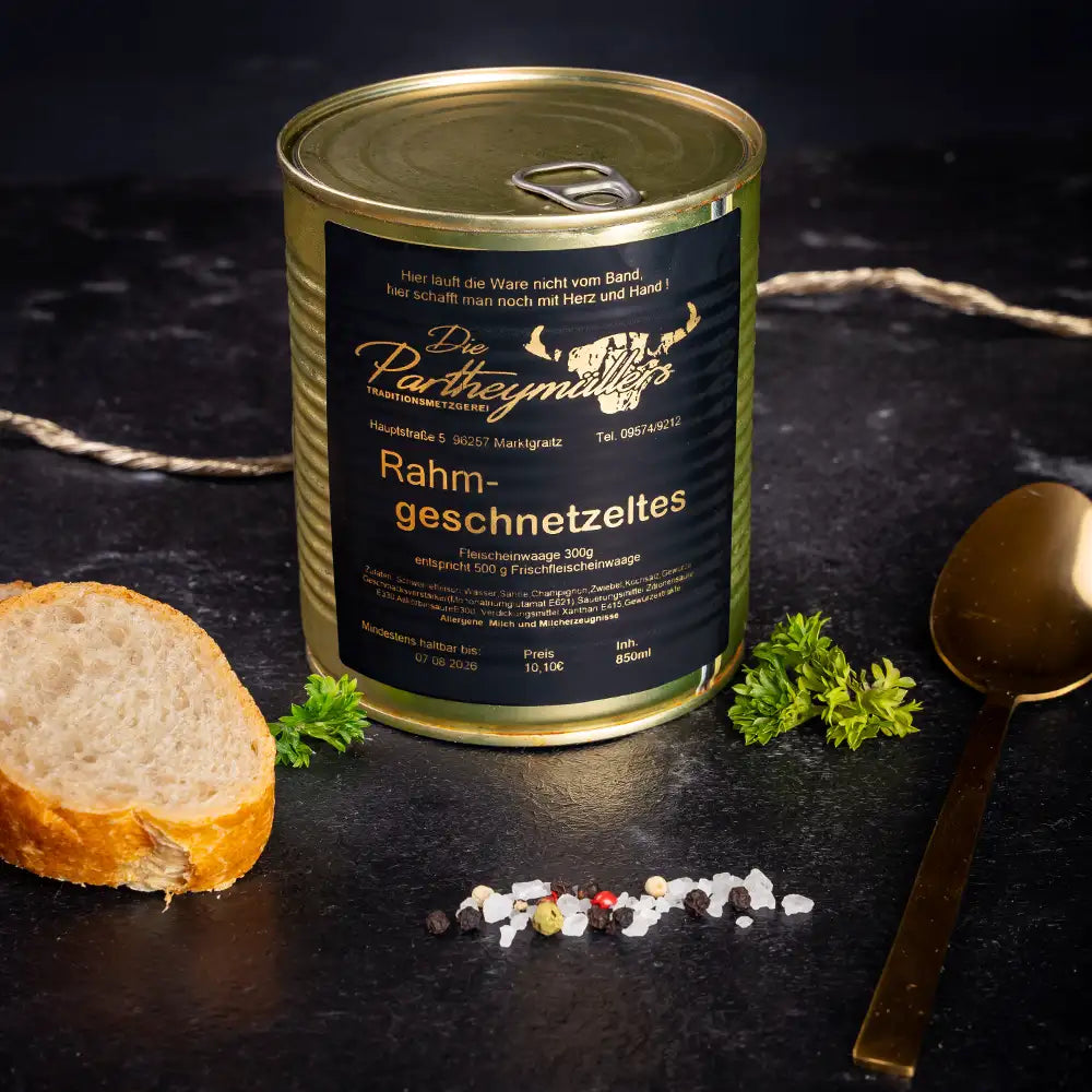 Goldene Konservendose der Metzgerei Partheymüller mit Rahmgeschnetzeltem edel präsentiert auf dunklem Untergrund mit goldenem Löffel, frischen Kräutern, Pfefferkörnern, Meersalz und einer Baguettescheibe.