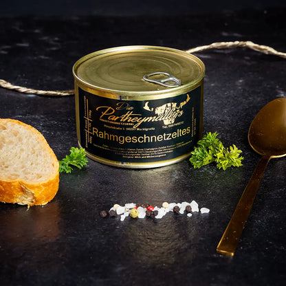 kleine goldene Konservendose der Metzgerei Partheymüller mit Rahmgeschnetzeltem edel präsentiert auf dunklem Untergrund mit goldenem Löffel, frischen Kräutern, Pfefferkörnern, Meersalz und einer Baguettescheibe.