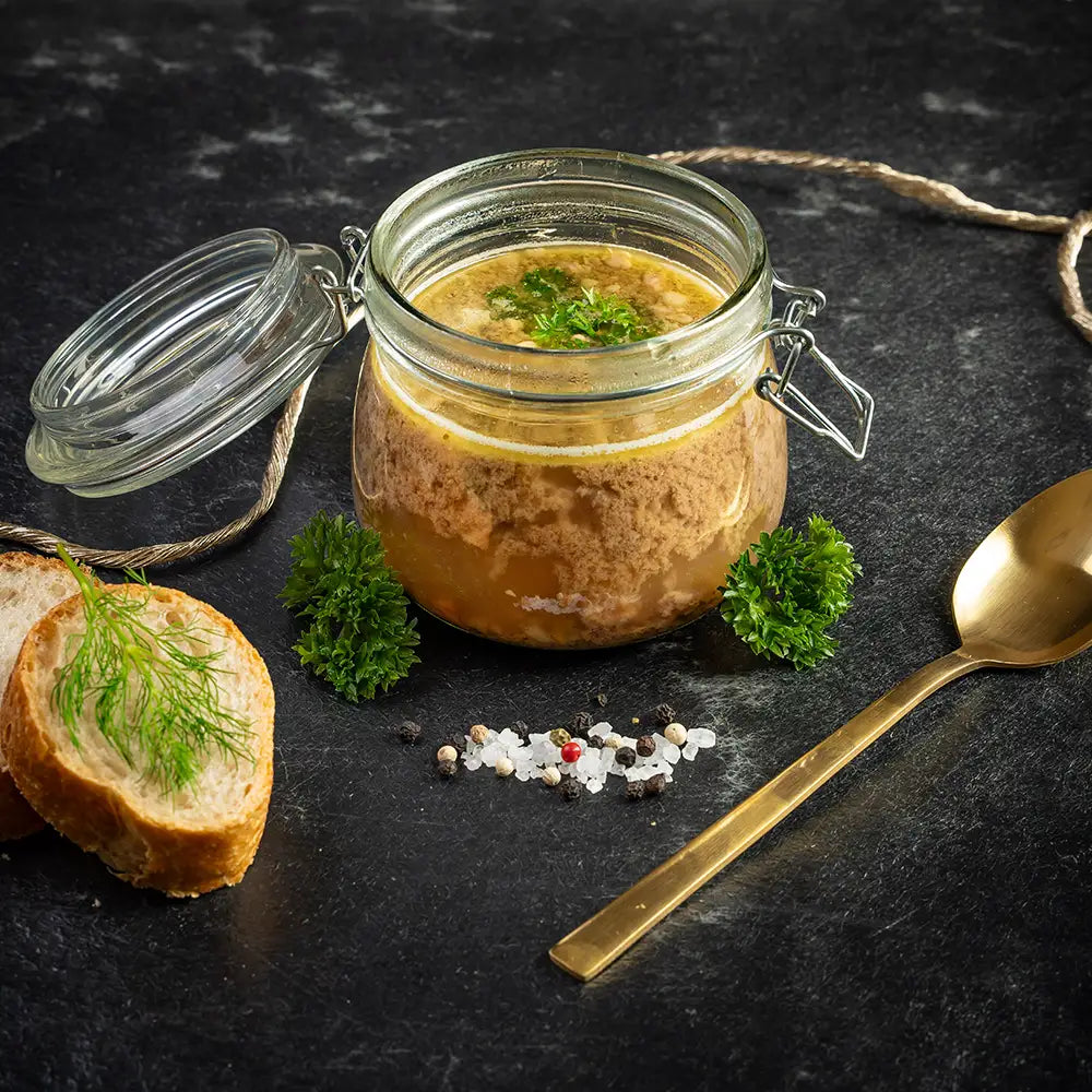 Rinderkraftbrühe im geöffneten Einmachglas mit Petersilie, daneben zwei Brotscheiben mit Dill, ein goldener Löffel und Gewürze auf dunklem Untergrund.