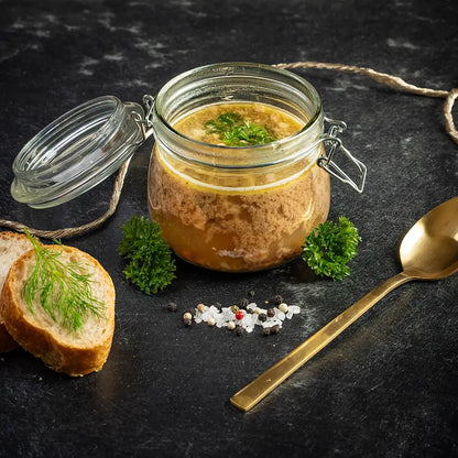 Rinderkraftbrühe im geöffneten Einmachglas mit Petersilie, daneben zwei Brotscheiben mit Dill, ein goldener Löffel und Gewürze auf dunklem Untergrund.