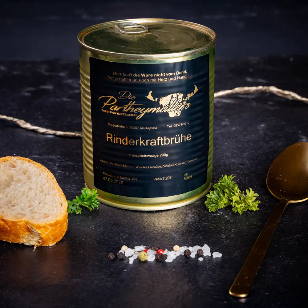 Goldene Konservendose der Metzgerei Partheymüller mit Rinderkraftbrühe edel präsentiert auf dunklem Untergrund mit goldenem Löffel, frischen Kräutern, Pfefferkörnern, Meersalz und einer Baguettescheibe.