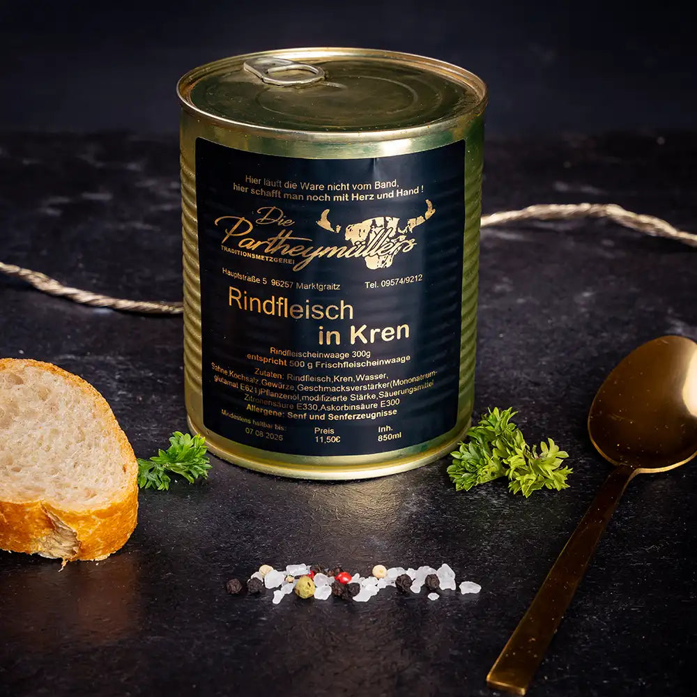 Goldene Konservendose der Metzgerei Partheymüller mit Rindfleisch in Kren edel präsentiert auf dunklem Untergrund mit goldenem Löffel, frischen Kräutern, Pfefferkörnern, Meersalz und einer Baguettescheibe.