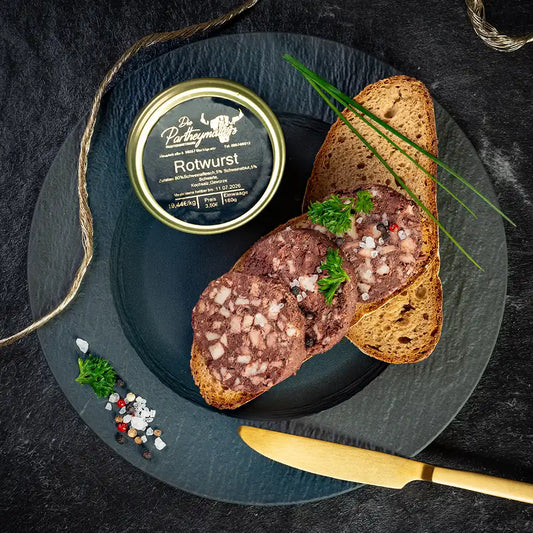 Drei Scheiben Rotwurst auf Bauernbrot, garniert mit Petersilie, grobem Salz und Pfefferkörnern, daneben ein Glas „Rotwurst“ von Partheymüller, frischer Schnittlauch und ein goldfarbenes Messer auf schwarzem Teller und dunklem Untergrund.