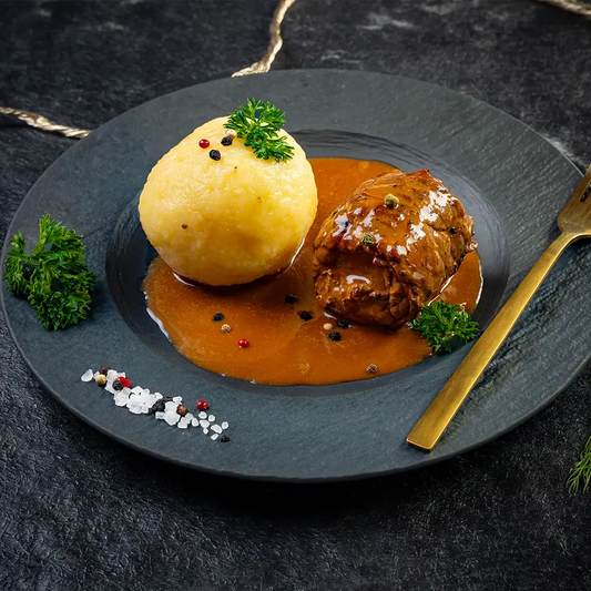 Rinderroulade mit Bratensauce und Kartoffelknödel auf schwarzem Teller, garniert mit Petersilie, Pfefferkörnern und grobem Salz, rechts eine goldene Gabel, im Hintergrund eine Kordel und Dillzweige auf dunklem Untergrund