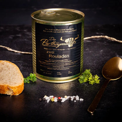 Goldene Konservendose der Metzgerei Partheymüller mit zwei Rouladen edel präsentiert auf dunklem Untergrund mit goldenem Löffel, frischen Kräutern, Pfefferkörnern, Meersalz und einer Baguettescheibe.