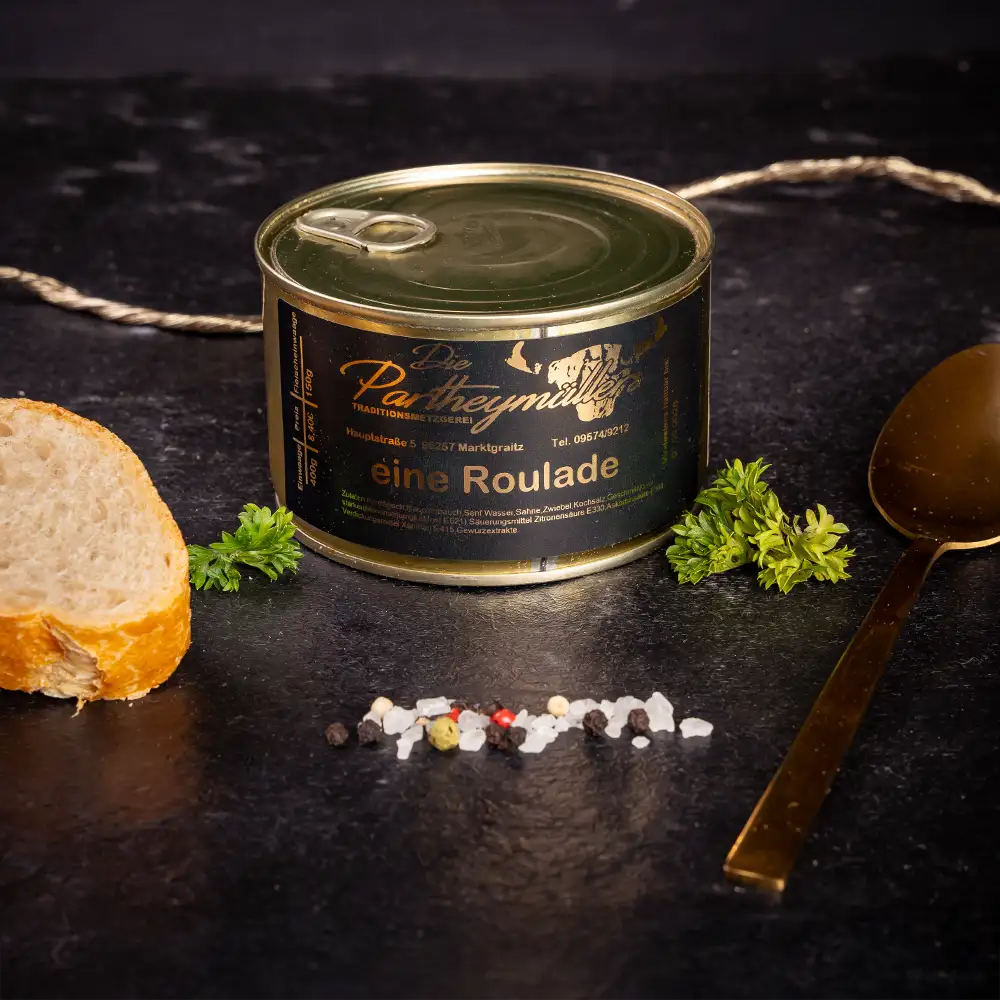 Goldene Konservendose der Metzgerei Partheymüller mit einer Roulade edel präsentiert auf dunklem Untergrund mit goldenem Löffel, frischen Kräutern, Pfefferkörnern, Meersalz und einer Baguettescheibe.