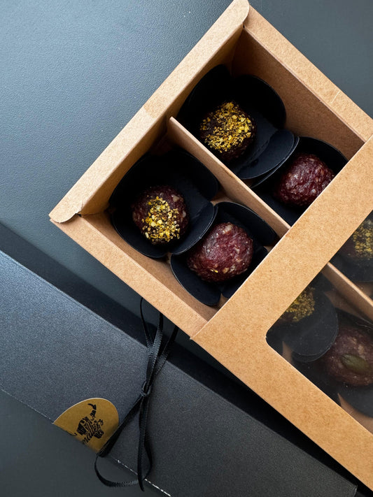 Geschenkverpackung mit  Salamipralinen in schwarzem Papier, teilweise mit Gewürzen bestreut, in einer offenen Kartonbox mit Sichtfenster, daneben der schwarze Deckel  mit goldfarbenem Logo