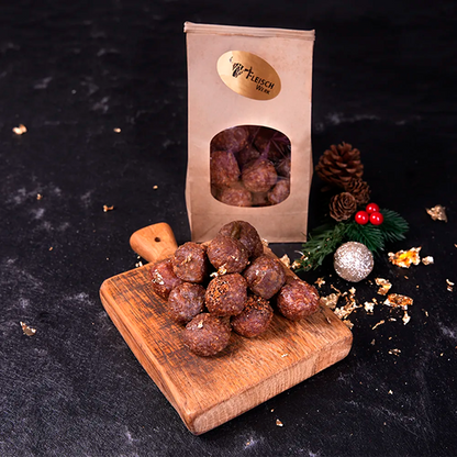 Eine kleine Pyramide aus Salamikugeln auf einem Holzbrettchen. Dahinter ein kartonfarbenes Tütchen mit Salamikugeln vor dunklem Hintergrund.