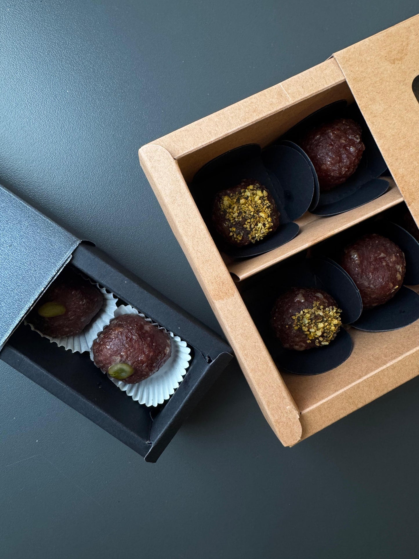 Zwei geöffnete Geschenkboxen mit Salamipralinen, eine in schwarzer Schachtel mit Pralinen in weißen Papierförmchen, die andere in brauner Kartonverpackung mit Pralinen in schwarzem Papier, teilweise mit Gewürzen oder Pistazien garniert