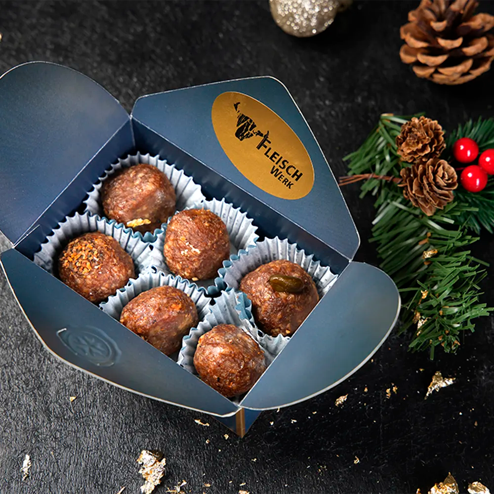 Sechs Salamikugeln in weißem Pralinenförmchen in blauer Schachtel vor dunklem Hintergrund.