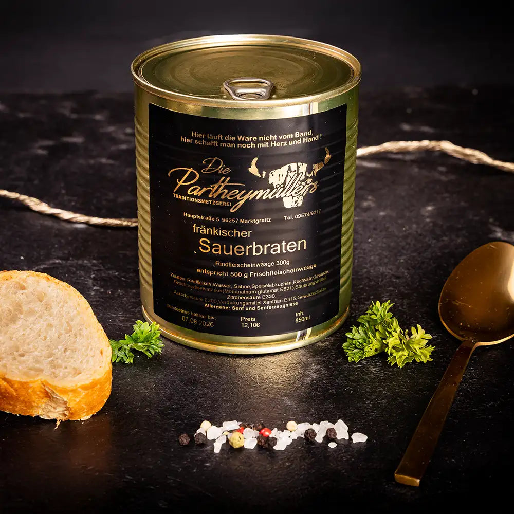 Goldene Konservendose der Metzgerei Partheymüller mit Sauerbraten edel präsentiert auf dunklem Untergrund mit goldenem Löffel, frischen Kräutern, Pfefferkörnern, Meersalz und einer Baguettescheibe.