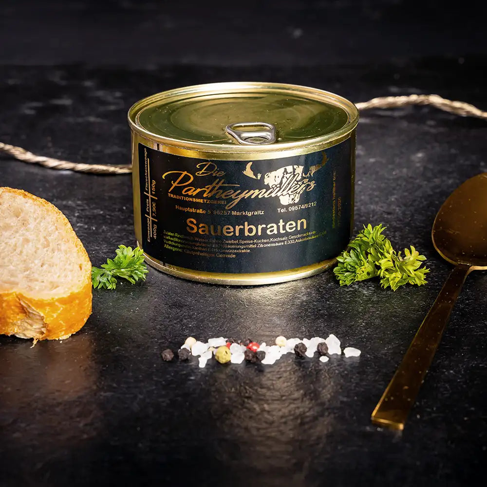 Kleine goldene Konservendose der Metzgerei Partheymüller mit Sauerbraten edel präsentiert auf dunklem Untergrund mit goldenem Löffel, frischen Kräutern, Pfefferkörnern, Meersalz und einer Baguettescheibe.