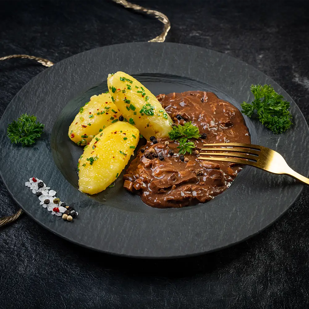 Saure Lunge in dunkler Sauce mit drei Petersilienkartoffeln auf schwarzem Teller, garniert mit Pfefferkörnern, Schnittlauch und frischer Petersilie, daneben eine goldene Gabel und dekorative Gewürze auf dunklem Untergrund.