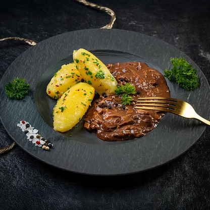Saure Lunge in dunkler Sauce mit drei Petersilienkartoffeln auf schwarzem Teller, garniert mit Pfefferkörnern, Schnittlauch und frischer Petersilie, daneben eine goldene Gabel und dekorative Gewürze auf dunklem Untergrund.
