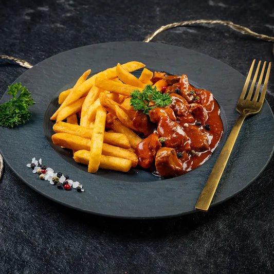 Schaschlik-Topf mit Fleischstücken in roter Sauce und einer Portion Pommes frites auf schwarzem Teller, garniert mit Petersilie, grobem Salz und Pfefferkörnern, daneben eine goldene Gabel auf dunklem Untergrund