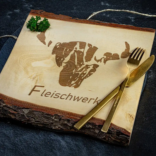 Dickes Schneidebrett mit Rindenkante, graviertem Logo eines Stierkopfes und dem Schriftzug „Fleischwerk“, daneben ein goldenes Besteck-Set aus Messer und Gabel sowie ein Zweig Petersilie.