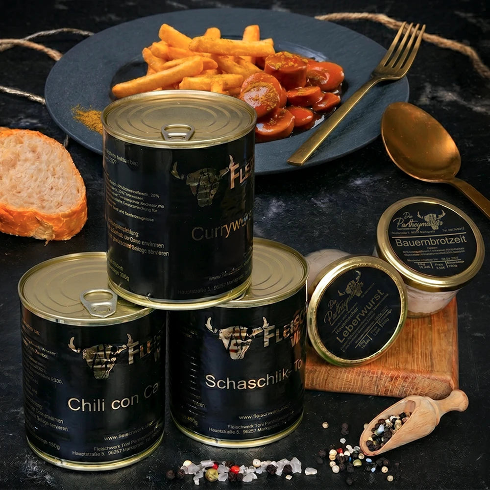 Drei Dosen mit dunklem Etikett und zwei Gläser mit dunklem Etikett stehen vor Teller mit Currywurst und Pommes.