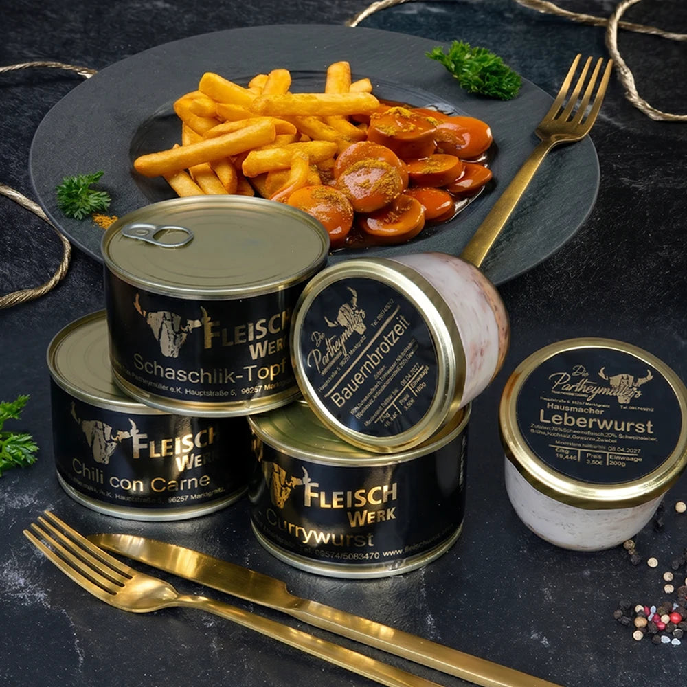 Drei Dosen mit dunklem Etikett und zwei Gläser mit dunklem Etikett stehen vor Teller mit Currywurst und Pommes.