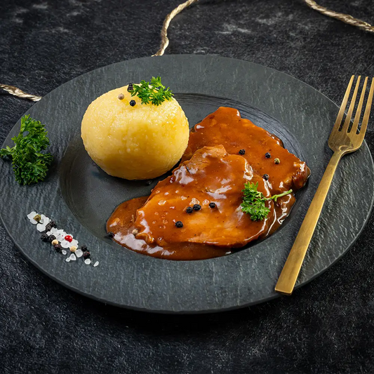 Schweinebraten mit zwei Fleischscheiben in dunkler Bratensauce und einem Kartoffelknödel auf schwarzem Teller, garniert mit Petersilie, Pfefferkörnern und grobem Salz, daneben eine goldene Gabel auf dunklem Untergrund.