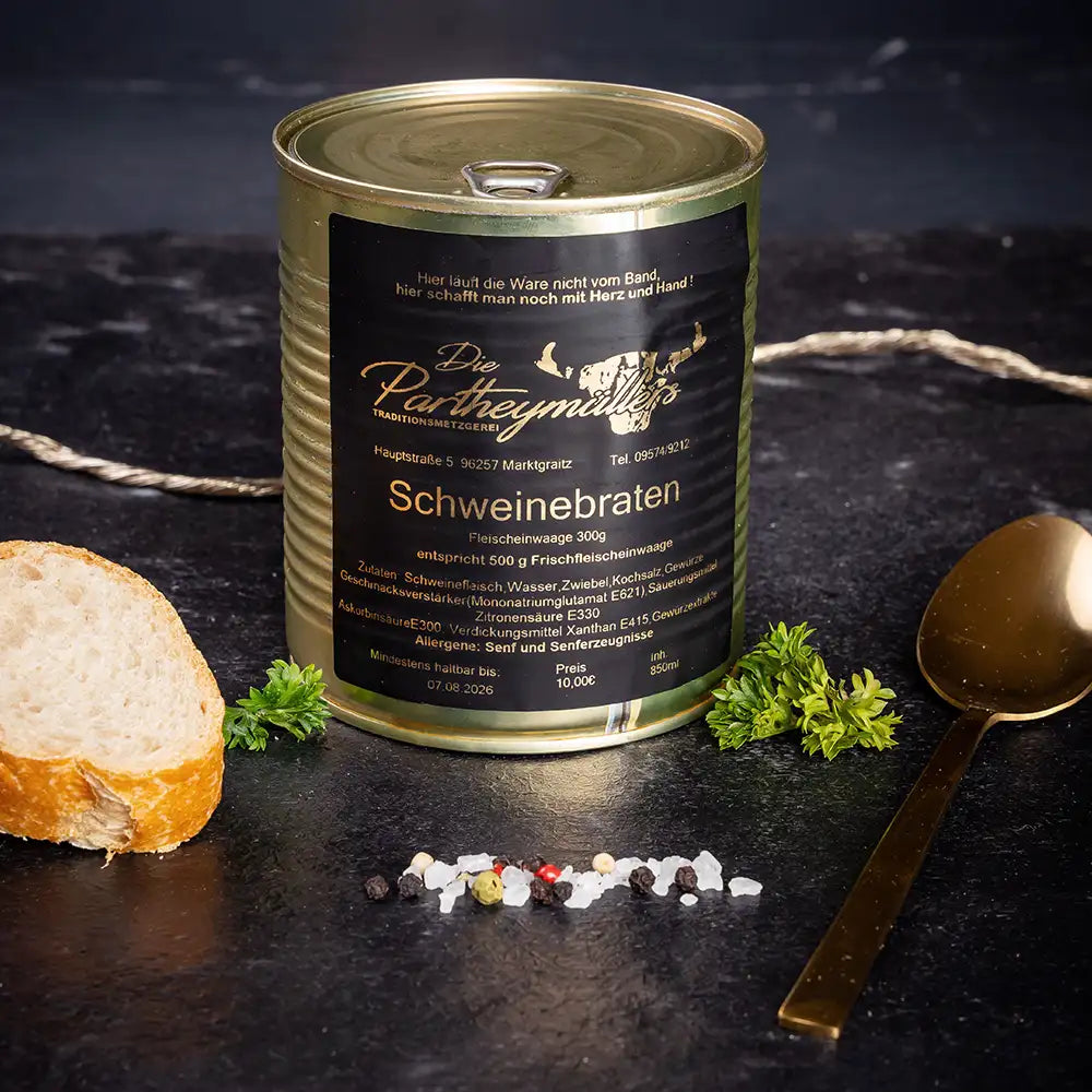 Goldene Konservendose der Metzgerei Partheymüller mit Schweinebraten edel präsentiert auf dunklem Untergrund mit goldenem Löffel, frischen Kräutern, Pfefferkörnern, Meersalz und einer Baguettescheibe