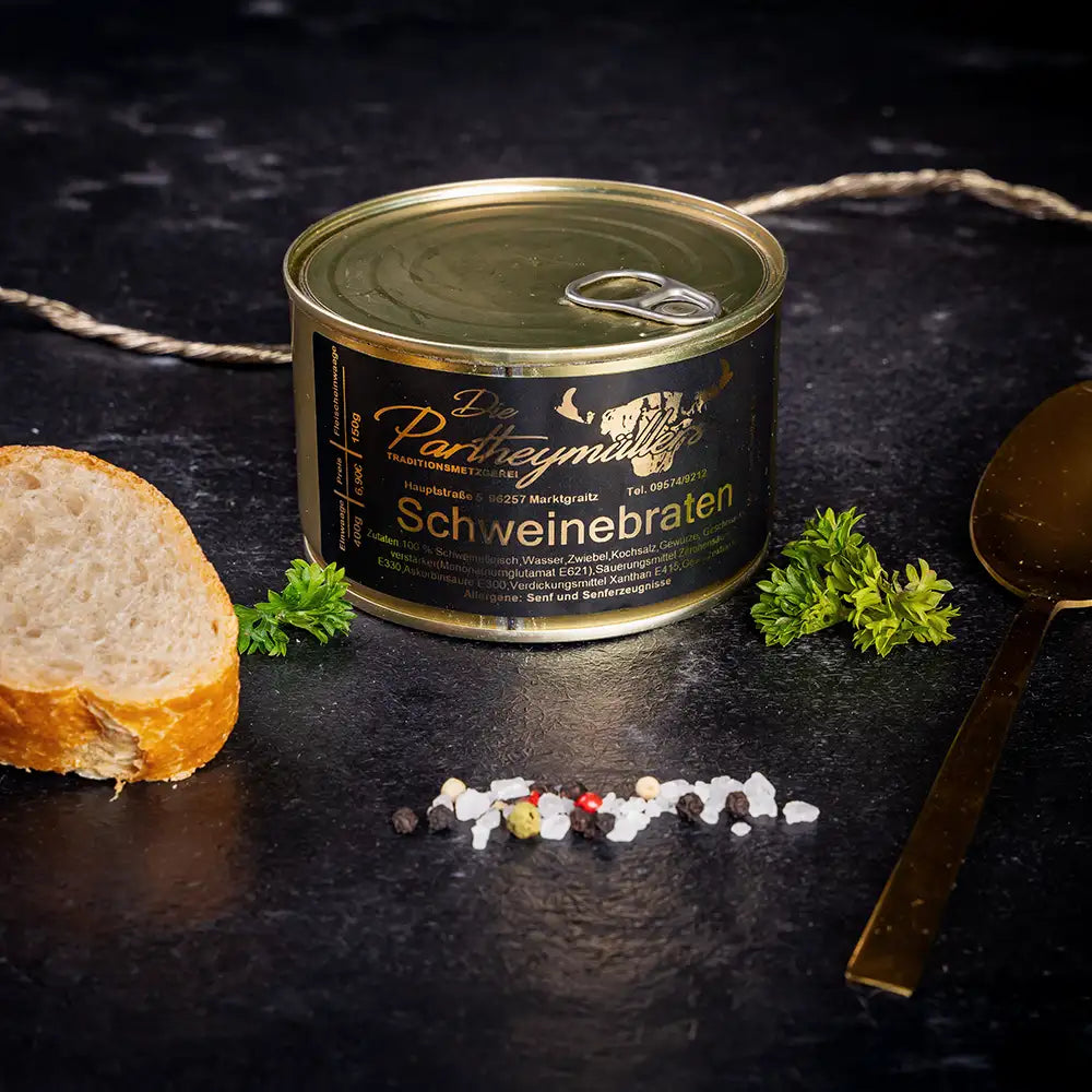 Kleine goldene Konservendose der Metzgerei Partheymüller mit Schweinebraten edel präsentiert auf dunklem Untergrund mit goldenem Löffel, frischen Kräutern, Pfefferkörnern, Meersalz und einer Baguettescheibe