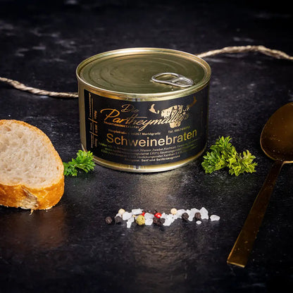 Kleine goldene Konservendose der Metzgerei Partheymüller mit Schweinebraten edel präsentiert auf dunklem Untergrund mit goldenem Löffel, frischen Kräutern, Pfefferkörnern, Meersalz und einer Baguettescheibe