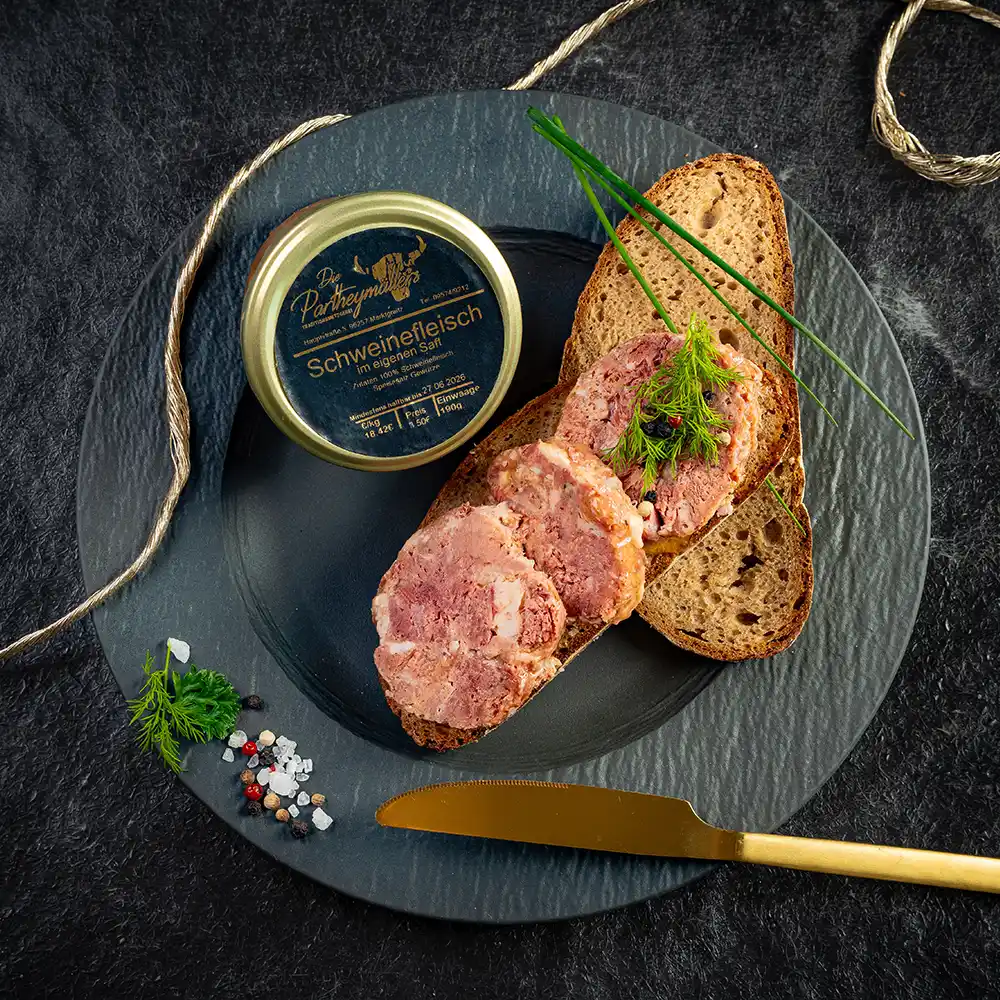 Drei Scheiben Schweinefleisch aus dem Glas auf Bauernbrot, daneben eine geöffnete Dose „Schweinefleisch im eigenen Saft“ von Partheymüller, angerichtet mit Schnittlauch, Dill, Pfefferkörnern und grobem Salz auf schwarzem Teller mit goldfarbenem Messer.