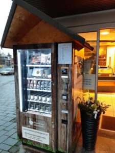 Wurstautomat mit Holzverkleidung und Satteldach vor einem Ladeneingang, befüllt mit Wurstgläsern und verpackten Fleischwaren, daneben ein schwarzer Blumentopf mit Blumendeko.