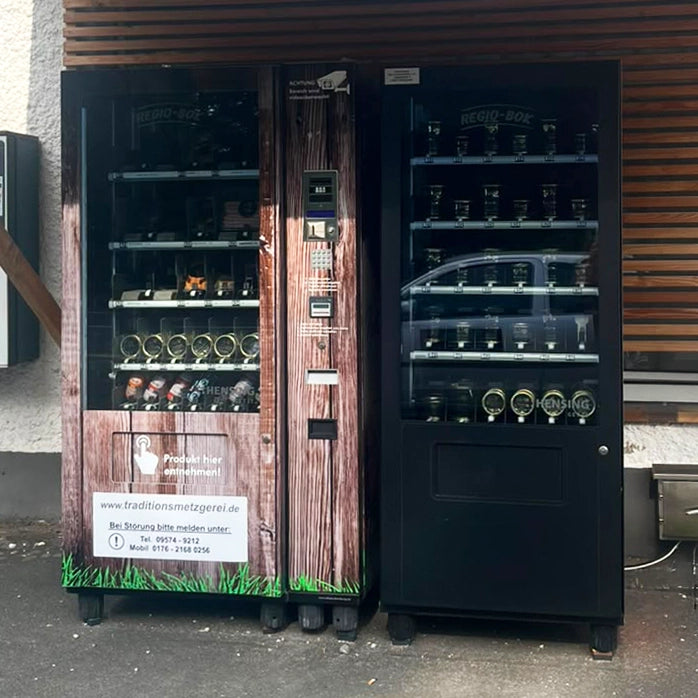 Ein Verkaufsautomat mit Holzoptik steht links neben einem schwarzen Verkaufsautomat. Beide haben ein transparentes Fenster in der Front, durch das man die Produkte sehen kann.