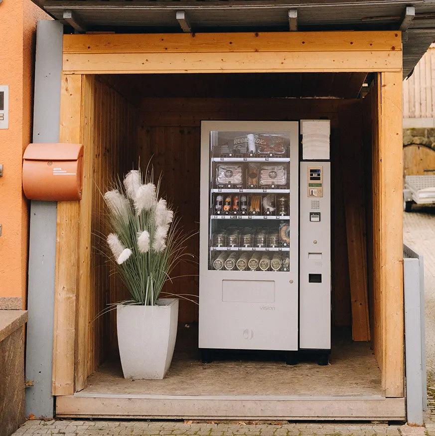 Verkaufsautomat mit regionalen Wurst- und Fleischprodukten in einem überdachten Holzunterstand, daneben ein Pflanzkübel mit Ziergras und ein orangefarbener Briefkasten an der Wand.
