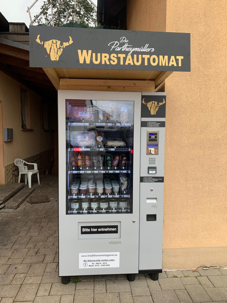 Wurstautomat der Metzgerei Partheymüller vor einer Hauswand, befüllt mit Gläsern, Dosen und vakuumverpackten Wurstwaren, mit Logo und Beschriftung „Wurstautomat“ auf dem Dachschild