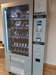 Grauer Wurstautomat im Innenraum auf Holzfußboden, befüllt mit Gläsern, verpackten Wurstwaren und Getränken, rechts Bedienfeld mit Display und Zahlungseinheit