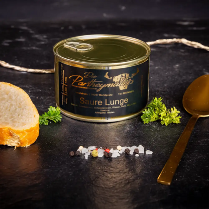 Kleine goldene Konservendose der Metzgerei Partheymüller mit saurer Lunge edel präsentiert auf dunklem Untergrund mit goldenem Löffel, frischen Kräutern, Pfefferkörnern, Meersalz und einer Baguettescheibe