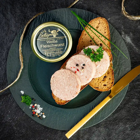 Drei Scheiben weiße Fleischwurst auf Bauernbrot, daneben ein Glas „Weiße Fleischwurst“ von Partheymüller, garniert mit Petersilie, Schnittlauch, Pfefferkörnern und grobem Salz, auf schwarzem Teller mit goldfarbenem Messer.